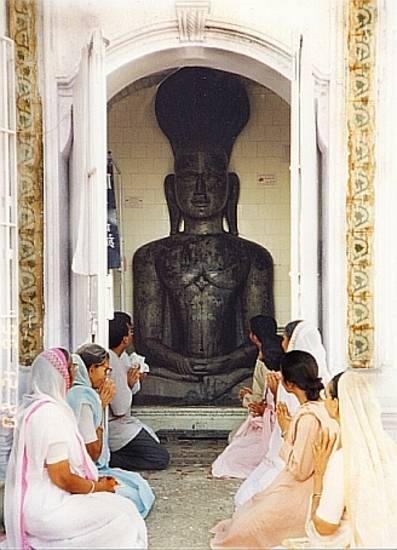 Lay men and women kneel in prayer before a large idol of a Jina in a temple. The idol's plain style and downcast eyes are characteristic of Digambara images.