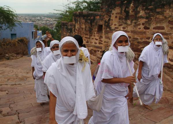 A group of Jain nuns walks barefoot up a hill. Dressed in white robes with their heads covered, they all wear cloths fixed over their mouths, attached by strings over the ears. This identifies them as either Śvetāmbara Sthānaka-vāsin or Terā-panthin nuns.