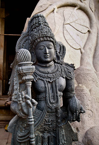 Carrying royal regalia, a statue of a goddess stands by the colossus of Bāhubali at Shravana Belgola. The nearly 18-metre-high statue of Bāhubali or Gommaṭeśvara – 'Lord of Gommaṭa' – has twin figures of goddesses either side