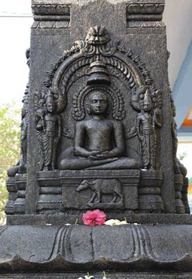 Carving of the 12th Jina, Vāsupūjya, with his buffalo emblem on the pedestal of a māna-stambha at Vilukkam in Tamil Nadu. This Digambara Jina is plainly sculpted though he meditates under an ornately carved arch. Recent offerings of flowers are below.